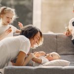 Side view of woman playing with toddler while other children standing nearby in living room